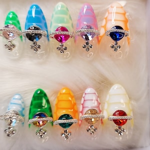 Può includere: Un set di 12 unghie finte colorate con un designo a strisce bianche e colorate. Ogni unghia ha un anello argentato con un centro gioiello.