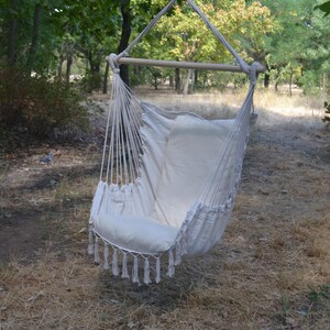 Puede incluir: Silla colgante de color crema con una barra de madera y soportes de cuerda. La silla tiene un asiento y respaldo acolchados, con borlas decorativas a lo largo del borde inferior. La silla est&aacute; suspendida de un &aacute;rbol en un entorno natural al aire libre.