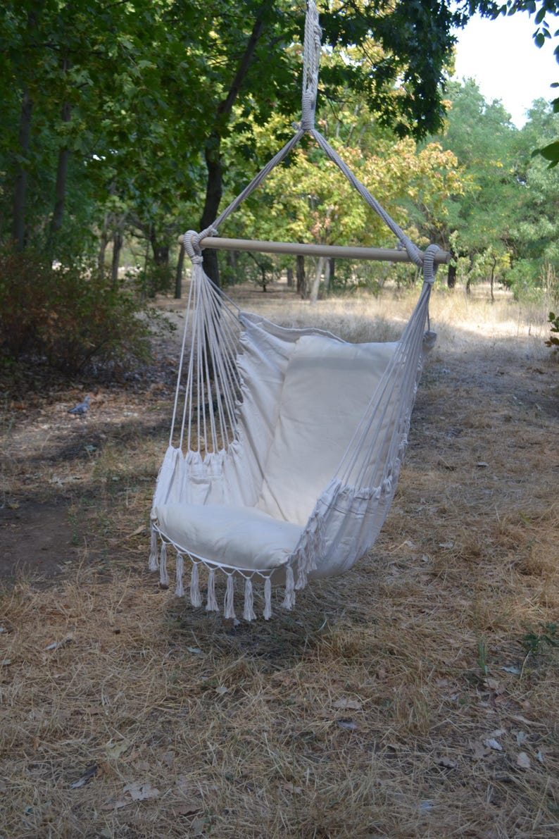 Puede incluir: Silla colgante de color crema con una barra de madera y soportes de cuerda. La silla tiene un asiento y respaldo acolchados, con borlas decorativas a lo largo del borde inferior. La silla est&aacute; suspendida de un &aacute;rbol en un entorno natural al aire libre.