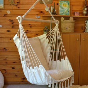 Puede incluir: Una silla hamaca de macram&eacute; blanco con una barra de madera y un coj&iacute;n color crema. La silla cuelga de una cuerda y tiene flecos en la parte inferior. El fondo es una pared de madera.