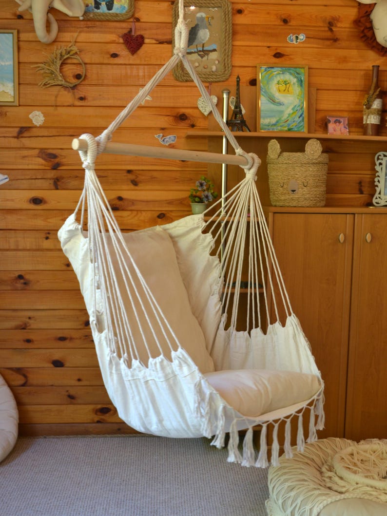 Puede incluir: Una silla hamaca de macram&eacute; blanco con una barra de madera y un coj&iacute;n color crema. La silla cuelga de una cuerda y tiene flecos en la parte inferior. El fondo es una pared de madera.