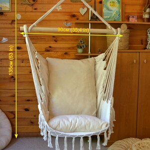 Puede incluir: Silla hamaca de macram&eacute; color crema con una barra de madera de 90 cm de ancho y una altura total de 135 cm. La silla tiene un coj&iacute;n grande y suave y borlas decorativas. Cuelga de una cuerda y est&aacute; colocada contra una pared de madera.