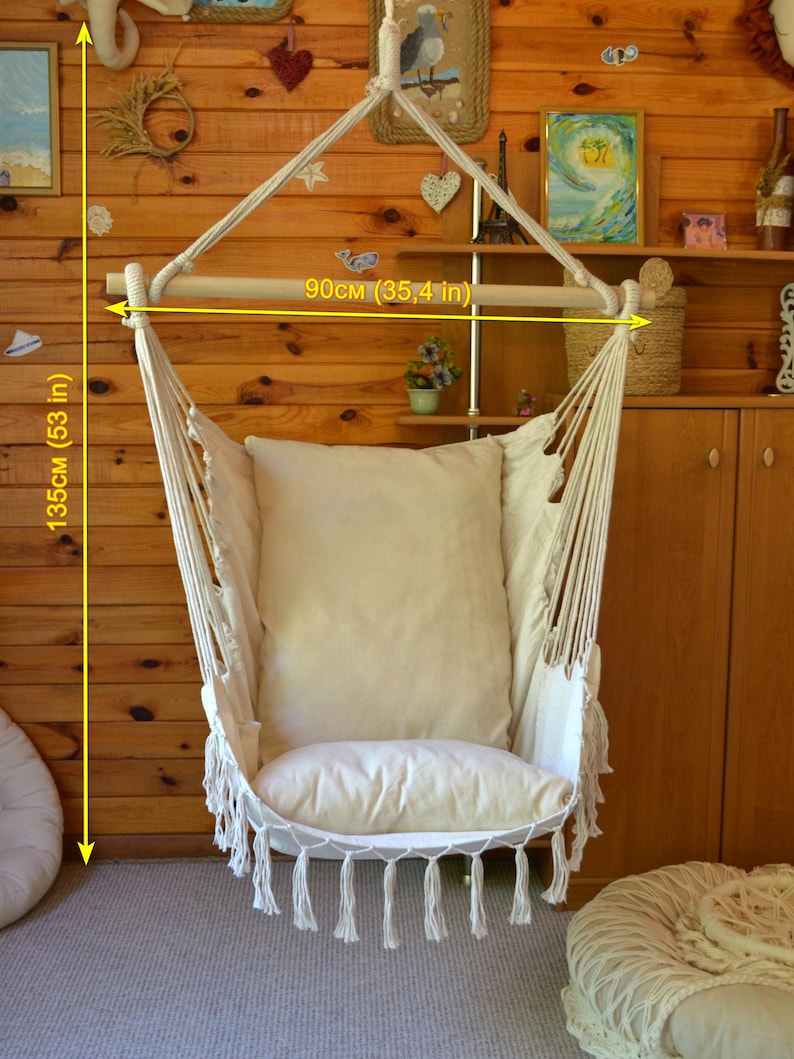 Puede incluir: Silla hamaca de macram&eacute; color crema con una barra de madera de 90 cm de ancho y una altura total de 135 cm. La silla tiene un coj&iacute;n grande y suave y borlas decorativas. Cuelga de una cuerda y est&aacute; colocada contra una pared de madera.