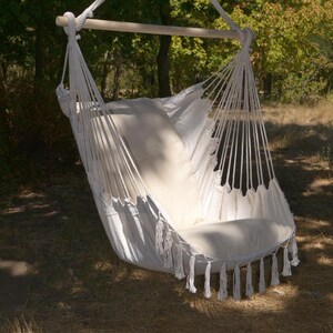 Puede incluir: Una silla hamaca de macram&eacute; blanco con un coj&iacute;n crema y borlas. La silla est&aacute; suspendida de una barra de madera y una cuerda, sobre un fondo de &aacute;rboles y luz solar, creando un espacio relajante al aire libre.