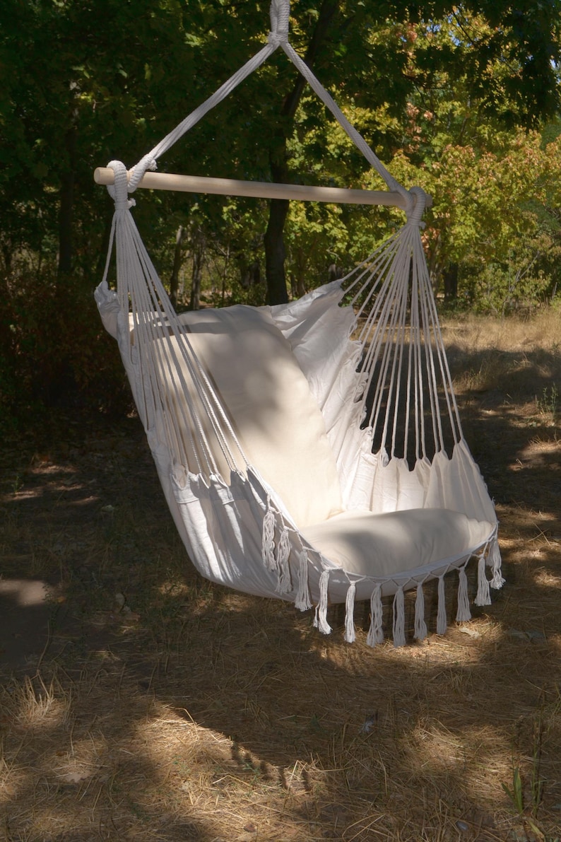 Puede incluir: Una silla hamaca de macram&eacute; blanco con un coj&iacute;n crema y borlas. La silla est&aacute; suspendida de una barra de madera y una cuerda, sobre un fondo de &aacute;rboles y luz solar, creando un espacio relajante al aire libre.