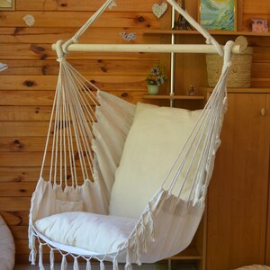 Puede incluir: Una silla colgante de tela y cuerda color crema, con una barra de madera en la parte superior. La silla tiene un coj&iacute;n grande y suave y un borde con flecos. Est&aacute; suspendida de una viga de madera, creando una zona de estar acogedora.