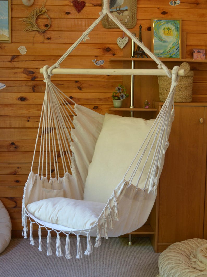 Puede incluir: Una silla colgante de tela y cuerda color crema, con una barra de madera en la parte superior. La silla tiene un coj&iacute;n grande y suave y un borde con flecos. Est&aacute; suspendida de una viga de madera, creando una zona de estar acogedora.