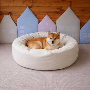 May include: A Shiba Inu dog rests in a round, off-white pet bed. The bed is on a light gray carpet, with colorful house-shaped wall decorations in the background. The dog has reddish-brown fur.