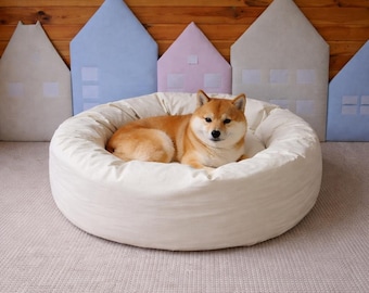 May include: A Shiba Inu dog lies in a circular, cream-coloured pet bed. The bed is on a light grey carpet, with colourful house-shaped wall decorations behind. The dog has reddish-brown fur.