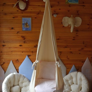 May include: A white fabric swing chair hanging from the ceiling in a room with wood paneling. The swing chair has a white cushion and a pink patterned fabric lining. There are two white cushions on the floor in front of the swing chair.