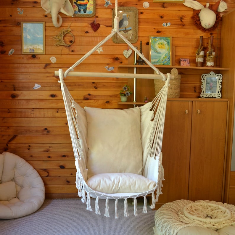 Puede incluir: Una silla colgante de color crema con una barra de madera y un coj&iacute;n grande y suave. La silla est&aacute; suspendida de una cuerda y tiene flecos en la parte inferior. El fondo es una pared con paneles de madera con objetos decorativos.