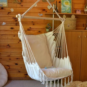 Puede incluir: Una silla colgante de tela blanquecina y cuerda, suspendida de una viga de madera. La silla tiene un coj&iacute;n grande y suave y flecos decorativos en el borde inferior. El fondo presenta paneles de madera y objetos decorativos.