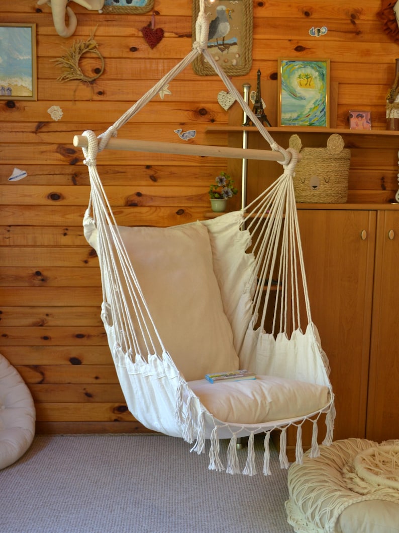 Puede incluir: Una silla colgante de tela blanquecina y cuerda, suspendida de una viga de madera. La silla tiene un coj&iacute;n grande y suave y flecos decorativos en el borde inferior. El fondo presenta paneles de madera y objetos decorativos.