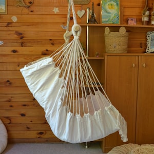 Puede incluir: Una hamaca de beb&eacute; de macram&eacute; blanco suspendida del techo. La hamaca est&aacute; hecha de tela y cuerda blancas, con un borde con flecos. El fondo es una pared de madera con objetos decorativos.