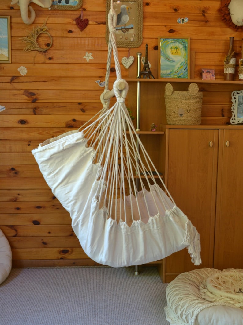 Puede incluir: Una hamaca de beb&eacute; de macram&eacute; blanco suspendida del techo. La hamaca est&aacute; hecha de tela y cuerda blancas, con un borde con flecos. El fondo es una pared de madera con objetos decorativos.