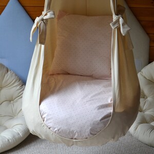 May include: A white fabric hanging chair with a pink and white patterned cushion. The chair is suspended from the ceiling and is ideal for relaxing or reading.