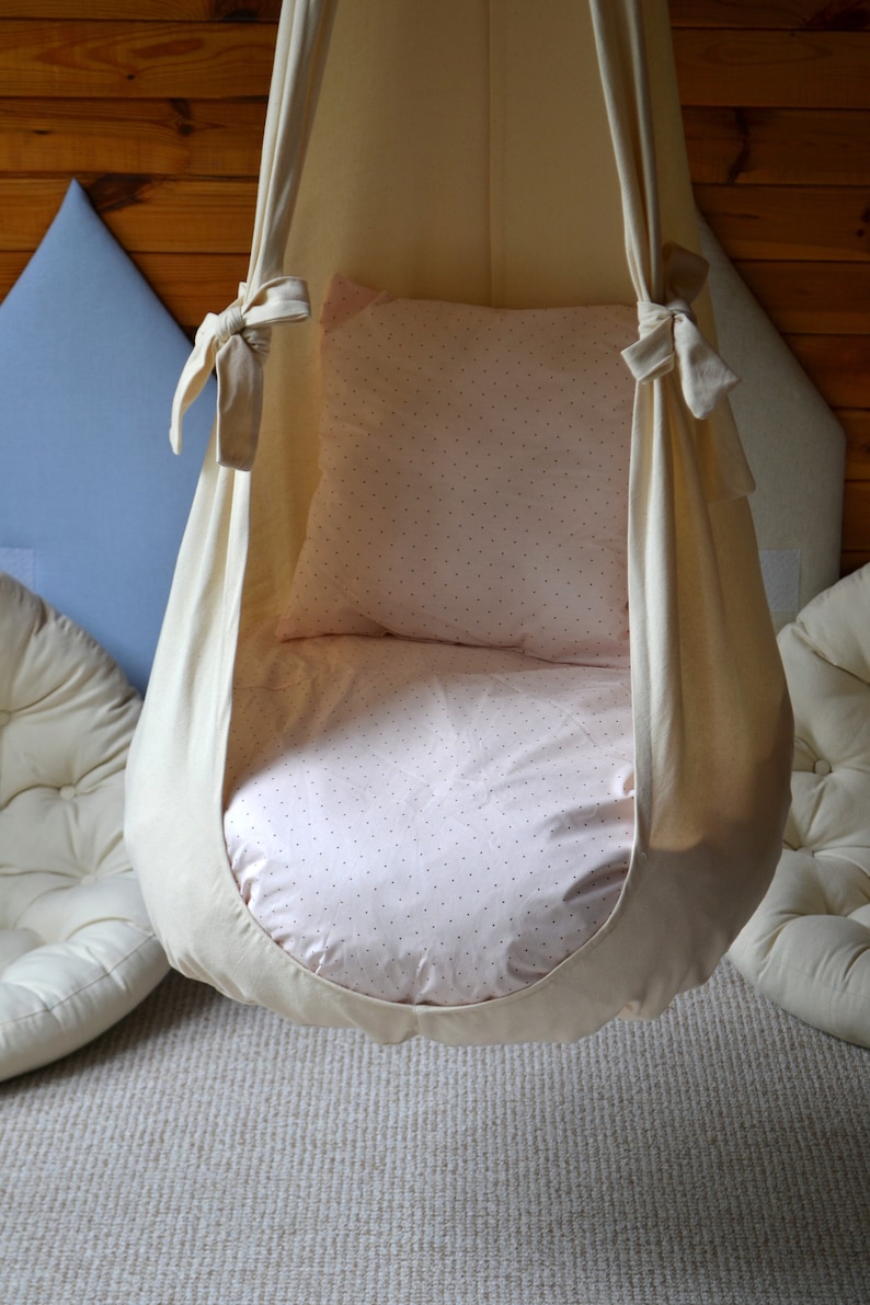 May include: A white fabric hanging chair with a pink and white patterned cushion. The chair is suspended from the ceiling and is ideal for relaxing or reading.