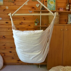 Puede incluir: Una silla hamaca de tela blanca suspendida de un marco de madera. La silla cuelga frente a una pared con paneles de madera, con objetos decorativos en estantes y la pared. La silla est&aacute; dise&ntilde;ada para la relajaci&oacute;n y el confort.