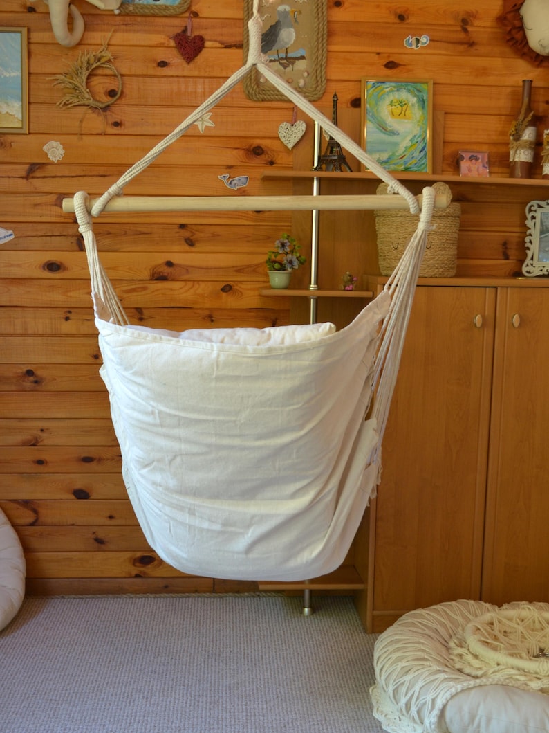 Puede incluir: Una silla hamaca de tela blanca suspendida de un marco de madera. La silla cuelga frente a una pared con paneles de madera, con objetos decorativos en estantes y la pared. La silla est&aacute; dise&ntilde;ada para la relajaci&oacute;n y el confort.