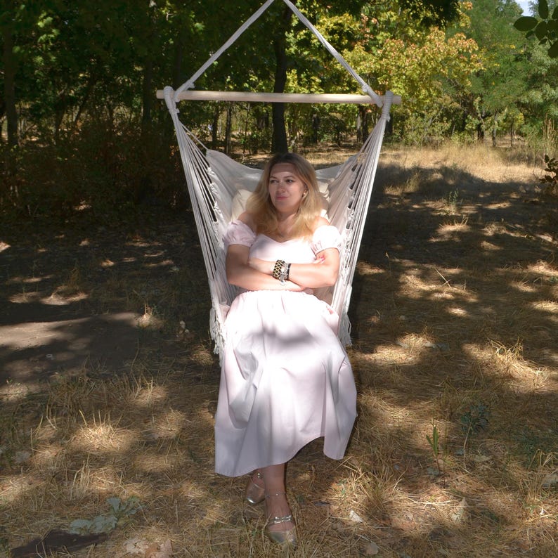 Puede incluir: Una silla hamaca blanca suspendida de un marco de madera y una cuerda, al aire libre. La silla tiene un asiento de tela blanca. El fondo incluye &aacute;rboles y luz solar. Una persona est&aacute; sentada en la silla hamaca.
