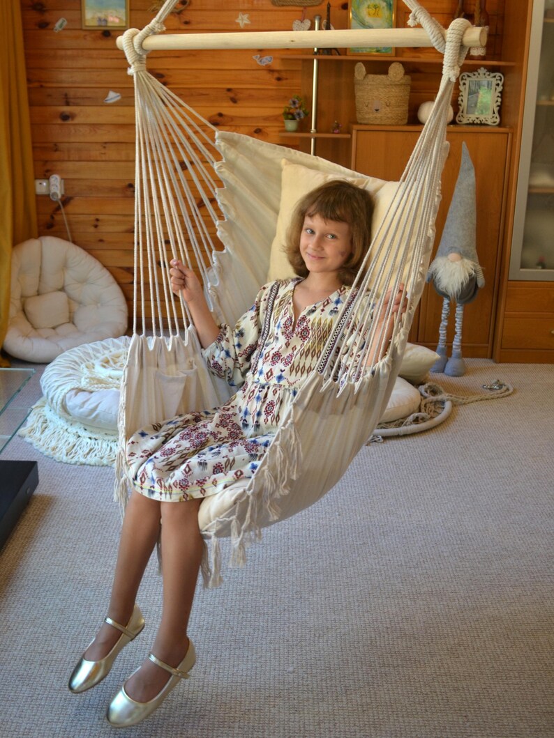Puede incluir: Una silla hamaca de macram&eacute; color crema con borde con flecos. La silla est&aacute; suspendida de una viga de madera y una cuerda. La persona en la silla lleva un vestido estampado y zapatos dorados. El fondo incluye paneles de madera y una alfombra neutra.