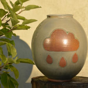May include: A ceramic vase with a light gray glaze and a brown cloud design with raindrops. The vase is sitting on a wooden surface.
