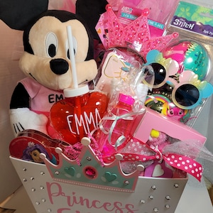 May include: A gift basket filled with princess-themed items. Includes a Mickey Mouse plush, a heart-shaped water bottle with "EMMA" on it, sunglasses, a crown, and other accessories. The basket is white with the words "Princess EMMA".