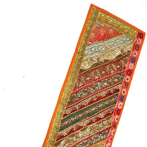 May include: A long, rectangular table runner with a colorful patchwork design. The runner is made of various fabrics and features intricate embroidery, sequins, and beads. The colors include orange, red, green, gold, and silver.