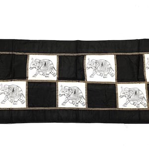 May include: Black and white table runner with a checkered pattern featuring embroidered elephants. The runner has a brown border.