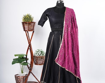 May include: A black ensemble on a mannequin, featuring a crop top, long skirt, and a draped magenta scarf. The skirt has gold horizontal stripes near the hem. The scarf has gold trim. A decorative plant stand with three baskets is in the background.