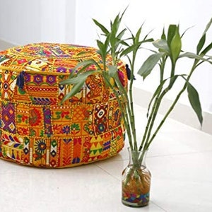 Può includere: Un pouf rotondo e colorato in patchwork con bordo giallo e nappe. Il pouf presenta vari motivi geometrici e floreali nei toni dell'arancione, giallo, viola e verde. Un vaso di vetro con steli di bambù verdi è accanto.