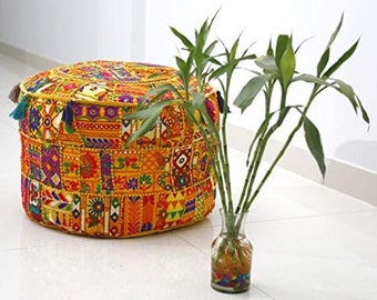 May include: A vibrant, patchwork pouffe with a yellow trim and tassels. The pouffe features a mix of geometric and floral designs in orange, yellow, purple, and green. A glass vase with bamboo stalks is placed next to it.