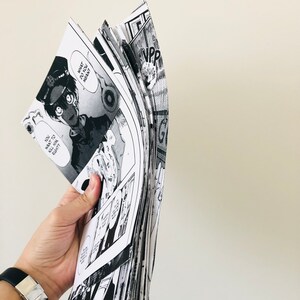 Peut inclure: Une pile de mangas en noir et blanc, tenue par une main. Les pages sont d&eacute;ploy&eacute;es, r&eacute;v&eacute;lant des cases avec des bulles de dialogue et des illustrations d&eacute;taill&eacute;es. Le texte "WHAT DO YOU MEAN?" est visible.