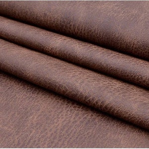 May include: Close-up of brown faux leather fabric with a textured surface. The fabric is folded in layers, showing the texture and color.