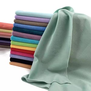Puede incluir: Una pila de 18 muestras de tela de diferentes colores, con una sola muestra más grande de tela verde claro doblada delante de la pila.
