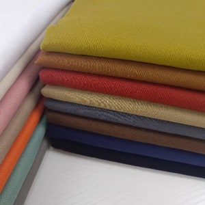 Puede incluir: Una pila de muestras de tela en varios colores, incluyendo blanco, beige, rosa, naranja, verde, azul, marrón, rojo y amarillo. La tela es una sarga con una textura sutil.