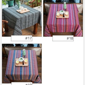 Op de afbeelding: Drie verschillende gestreepte tafelkleden in zwart-wit, roze en paars, en blauw en rood. Elk tafelkleed wordt getoond op een houten tafel met een theepot en theekopjes op een houten dienblad.