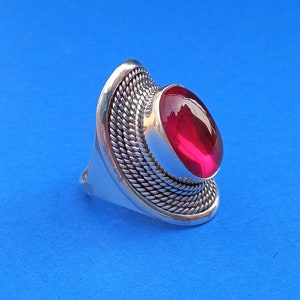 Może przedstawiać: Srebrny pierścionek z dużym, owalnym, jaskraworóżowym kamieniem szlachetnym osadzonym w środku. Obrączka pierścionka jest ozdobiona plecionym wzorem.