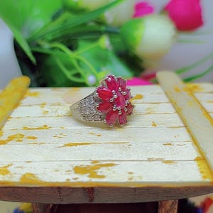 以下が含まれることがあります： 花の形に赤い宝石が並んだ銀の指輪。指輪には小さな白い宝石が飾られています。