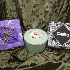 May include: Three decorative boxes with different designs. The first box is purple with a gold and purple dragonfly on the front. The second box is green with a floral design on the lid. The third box is black and white with a black square in the center and a white knot design.