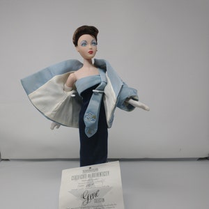 May include: A fashion doll wearing a strapless navy blue gown and a light blue jacket with white lining. The jacket has embroidered floral details. The doll has brown hair and is wearing white gloves. A certificate of authenticity is visible.