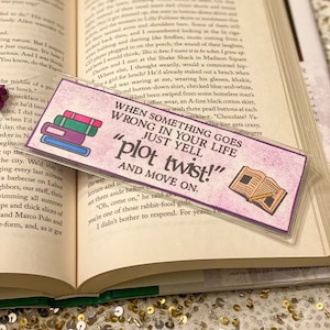 May include: Purple and pink bookmark with a stack of books and a pencil on an open book. The bookmark has the text "When something goes wrong in your life just yell "plot twist!" and move on."