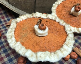 May include: Close-up of two decorative pumpkin pie ornaments. Each pie has an orange filling, a white crust, and a small pumpkin and cinnamon stick on top. The pies are on a plaid surface, with additional pumpkins and cinnamon sticks.