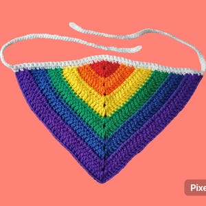 May include: A handmade crochet bandana in a rainbow color scheme, featuring red, orange, yellow, green, blue, and purple. The bandana has a white border and white ties for securing. The item is displayed against a coral background.