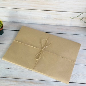 K&ouml;nnte beinhalten: Ein braunes Papierpaket, das mit Schnur auf einer wei&szlig;en Holzoberfl&auml;che gebunden ist. Im Hintergrund befinden sich zwei kleine Topfkakteen.