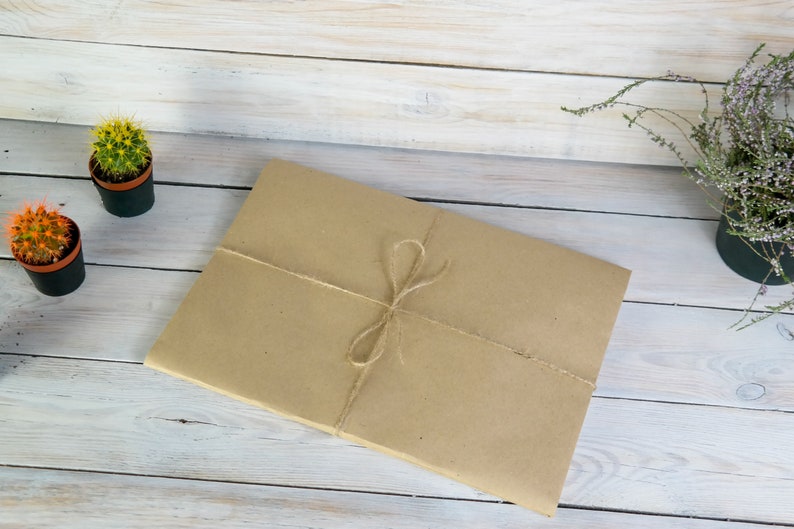 K&ouml;nnte beinhalten: Ein braunes Papierpaket, das mit Schnur auf einer wei&szlig;en Holzoberfl&auml;che gebunden ist. Im Hintergrund befinden sich zwei kleine Topfkakteen.