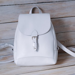 Puede incluir: Una mochila de cuero blanco con un cierre de hebilla simple y correas ajustables.