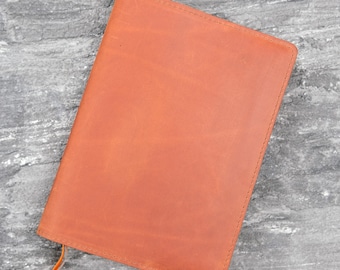 May include: A rectangular notebook cover made of smooth, tan-colored leather. The cover features visible stitching along the edges and a thin, matching leather ribbon bookmark extending from the bottom left corner of the front panel.