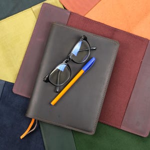 Puede incluir: Una funda de cuero gris oscuro para cuaderno con un par de gafas de montura negra y un bolígrafo naranja con tapa azul encima. El cuaderno está rodeado por varias otras fundas de cuero en colores amarillo, naranja y verde.