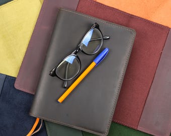 May include: A dark gray leather notebook cover featuring a pair of black-rimmed eyeglasses and an orange ballpoint pen with a blue cap resting on top. The notebook is surrounded by several other leather covers in yellow, orange, and green.